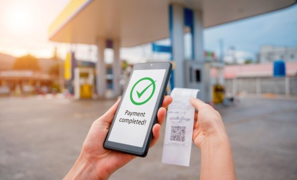 A Close-Up of a Smartphone Showing a "Payment Completed" Message, with a Printed Receipt and a Gas Station in the Background